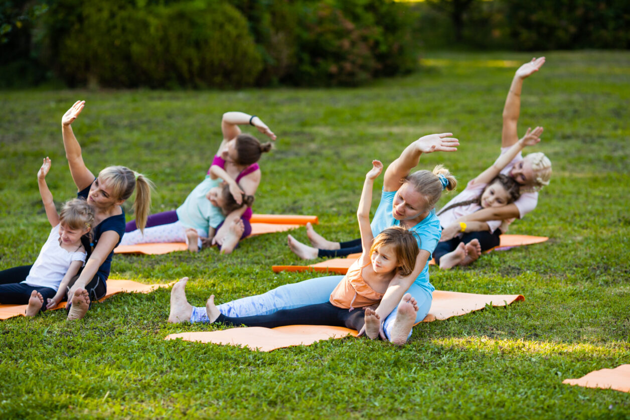 Group of mothers and their children doing exercise practicing yoga on a fitness rugs on the fresh air